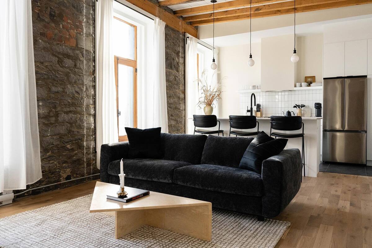 A modern living room with a dark grey sofa, a wooden coffee table, and a white rug. A kitchen with white cabinets and a stainless steel refrigerator is visible in the background.