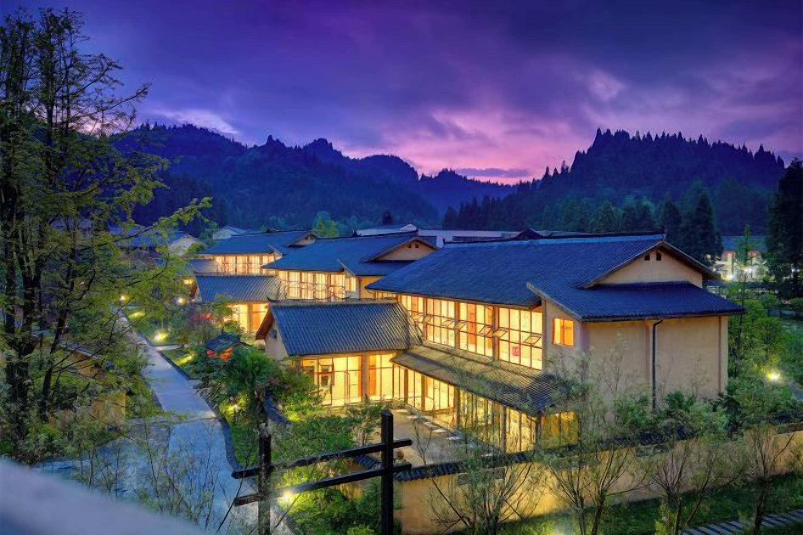 Un complejo de edificios tradicionales al estilo japonés, rodeado de vegetación y con montañas al fondo.