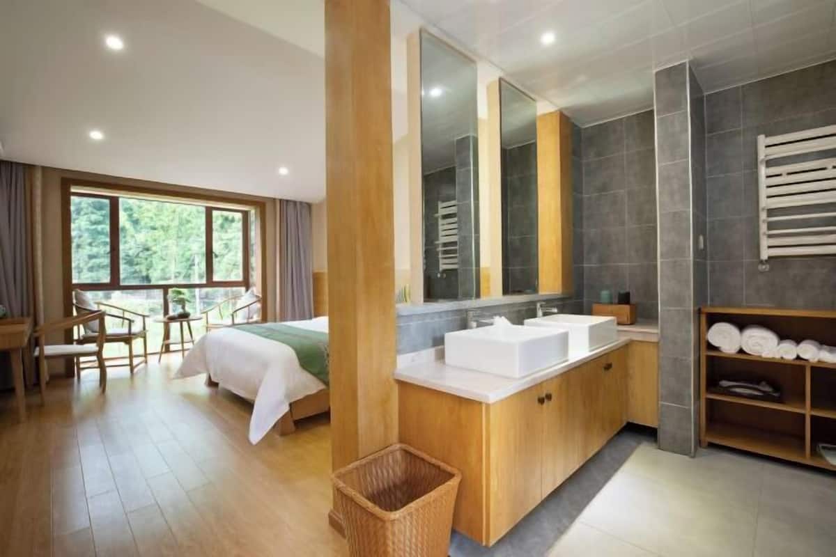 A modern hotel room with a large window, a wooden bed, a bathroom with a large mirror, and a wooden floor.
