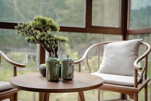 Una mesa de madera redonda con dos teteras verdes y un bonsái, una silla de mimbre con un cojín blanco, y vistas a la vegetación a través de una ventana.