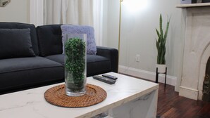 En stue med en sort sofa, en glass vase med grønne planter, en fjernbetjening og et hvidt marmorbord.