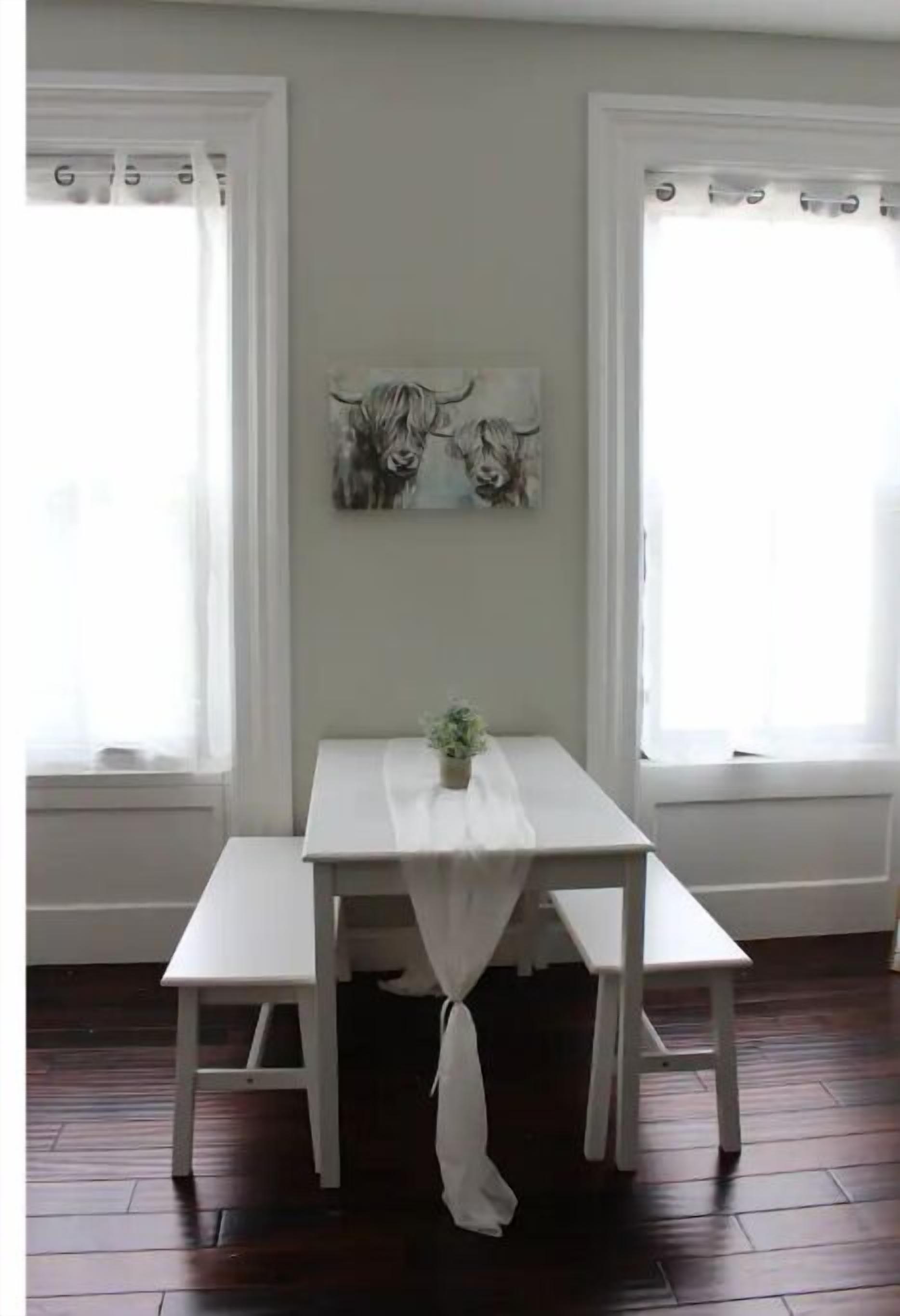 Un comedor con una mesa blanca y bancos, dos ventanas con cortinas y un cuadro en la pared.
