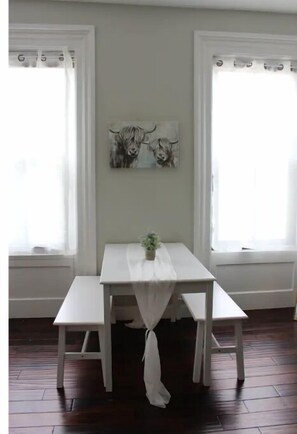 A dining area with a white table and benches, two windows with curtains, and a painting on the wall. - Chic, Renovated Brownstone in Prime DT Albany (Albany)