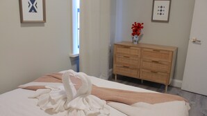 Un lit bien fait, avec une serviette et une couverture, une commode en bois ornée d’un vase rempli de fleurs rouges, et un tableau encadré accroché au mur.