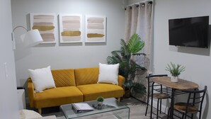 Un salon avec un canapé jaune, une table basse en verre, un téléviseur fixé au mur et une petite table en bois avec des chaises.