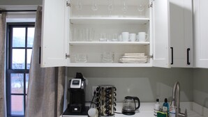 Une cuisine avec des meubles blancs, une machine à café, une bouilloire et divers articles en verre disposés sur des étagères.