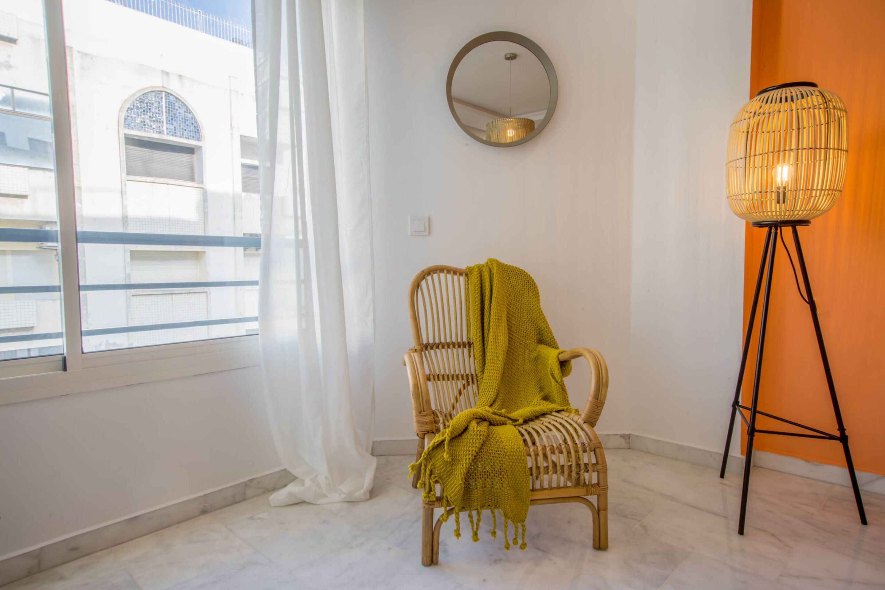 Ein Zimmer mit Korbsessel, Stehlampe, Spiegel an der Wand und einem Fenster mit Vorhängen.