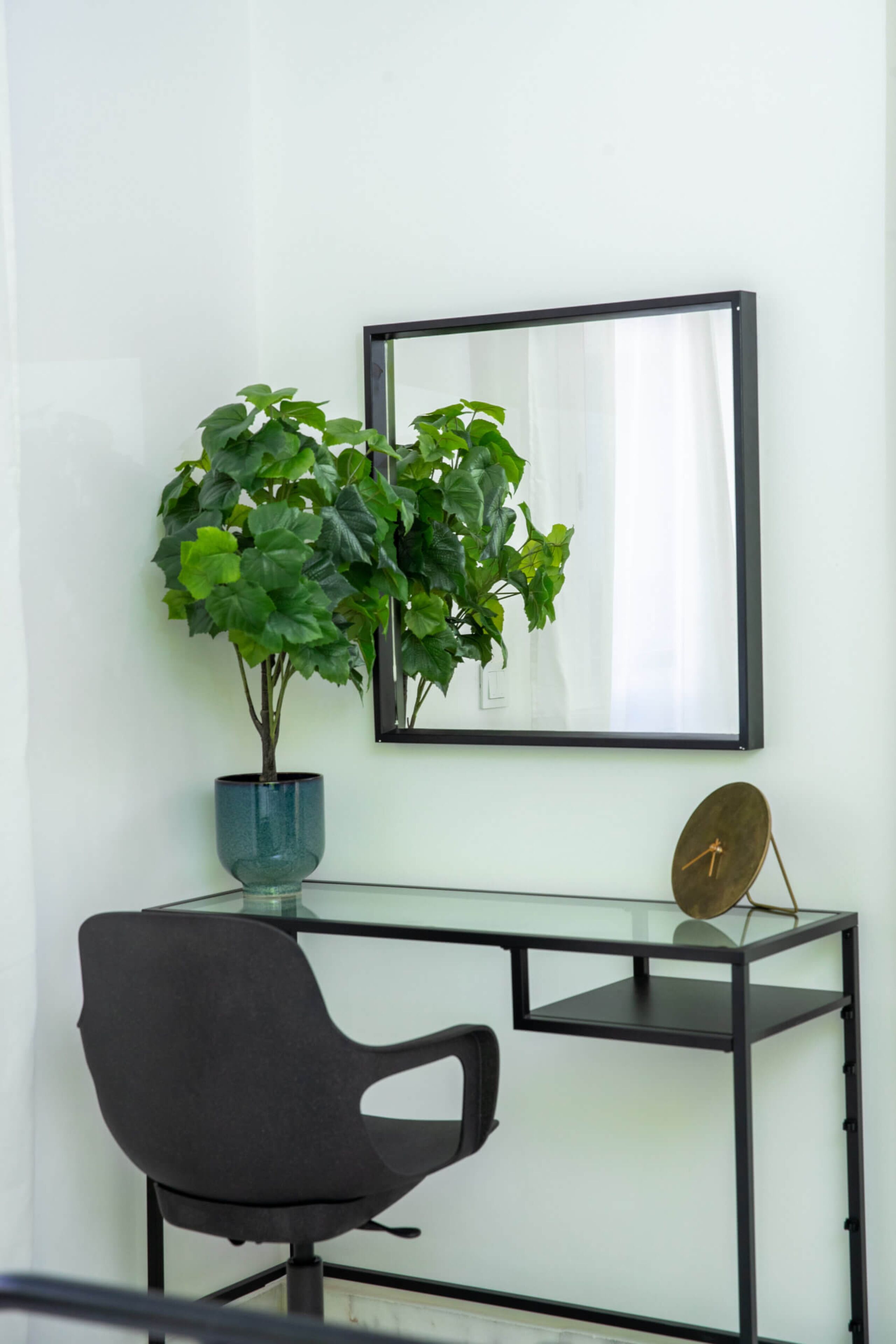 A desk with a chair, a potted plant, a mirror, and a clock on a glass table against a white wall.