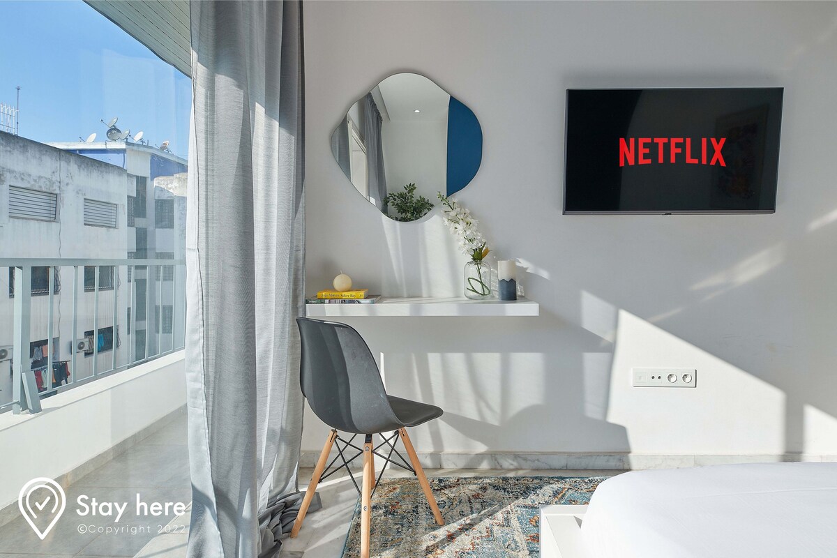 A modern hotel room with a large window, a round mirror, a flat-screen TV displaying 'NETFLIX', a black chair, a small shelf with decorative items, and a bed.