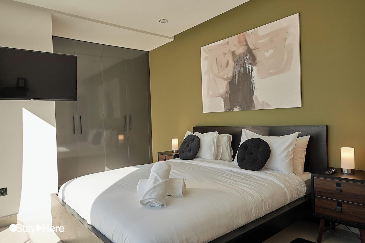 A modern bedroom with a large bed, a flat-screen TV, a painting on the wall, and a nightstand with a lamp.