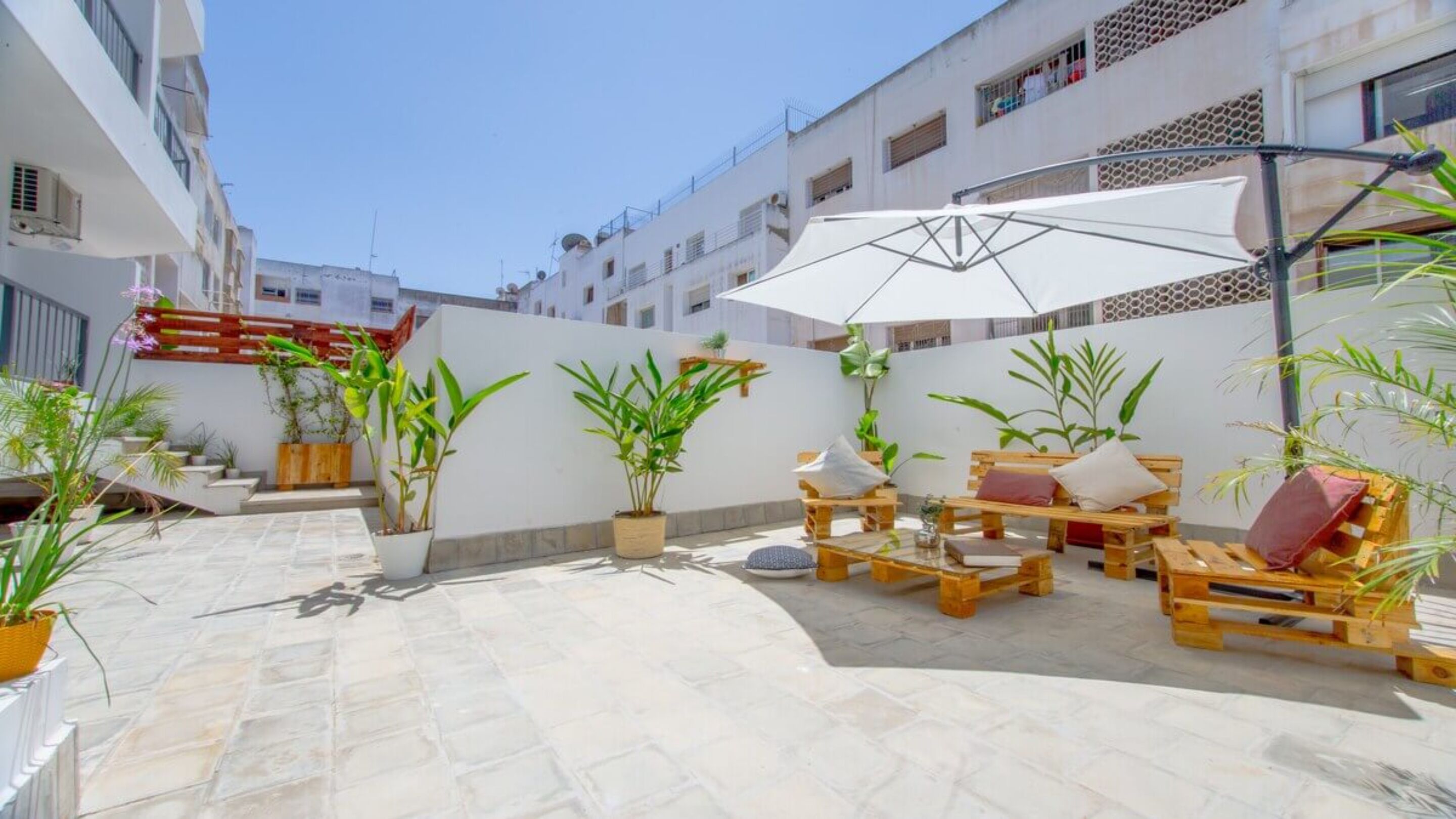 Un patio con mobiliario exterior, plantas en macetas y un paraguas blanco.