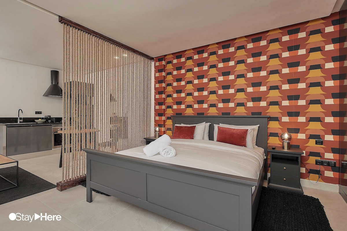 A modern bedroom with a grey bed, a patterned wall, a kitchen area, and a decorative curtain.