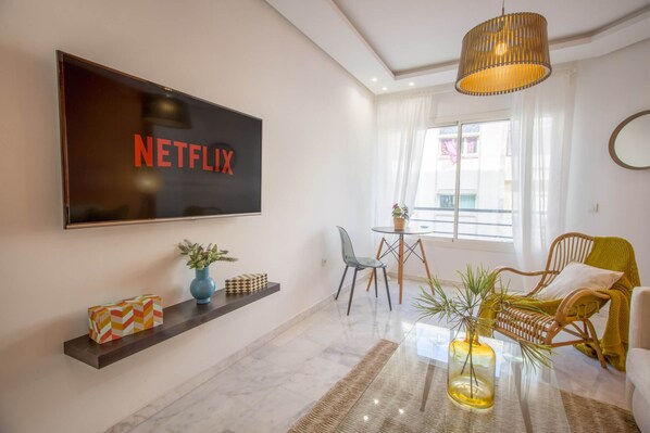 A modern living room with a large flat-screen TV mounted on the wall, a glass coffee table, wicker chairs, and a small side table with decorative items.