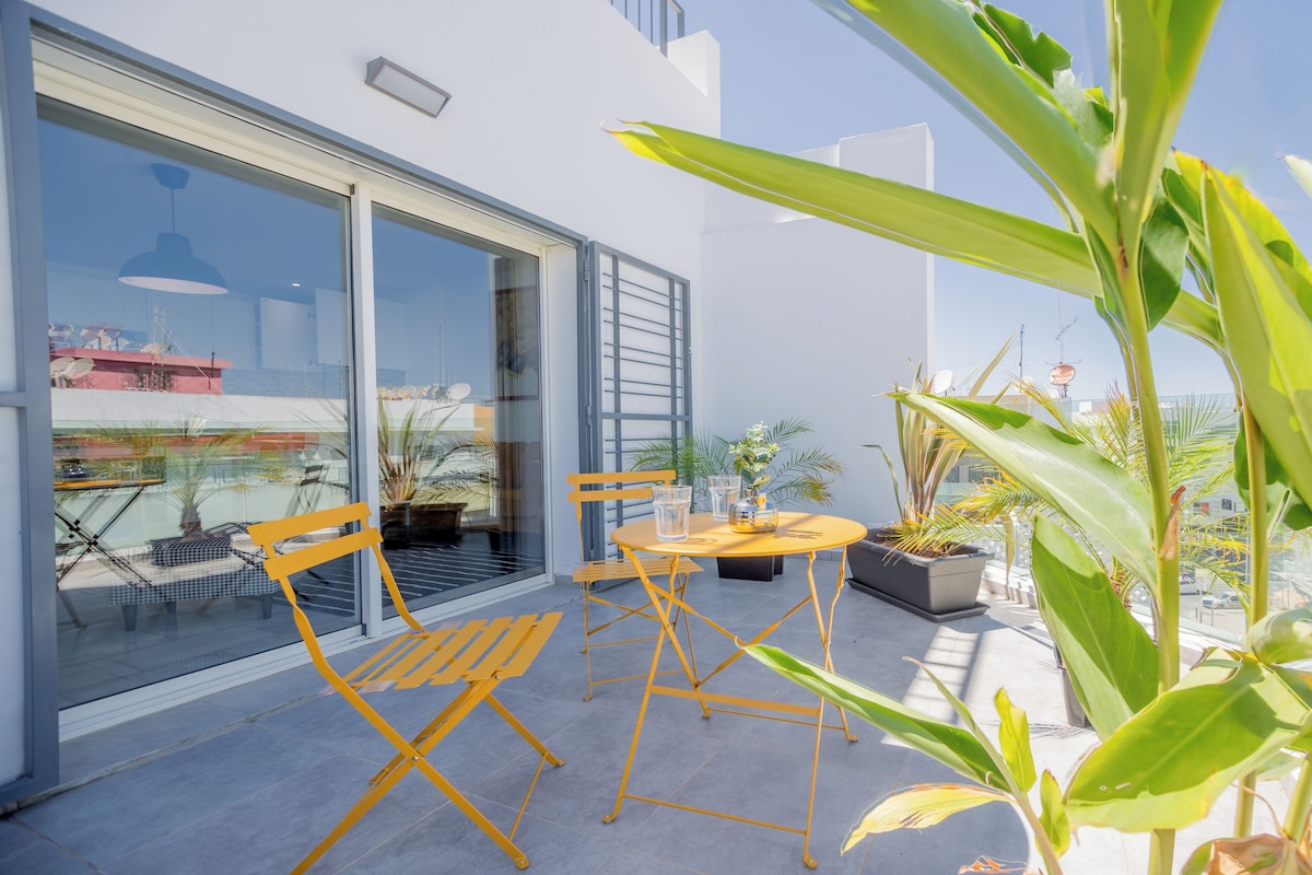A balcony with a glass door, a yellow table, and chairs, surrounded by potted plants.