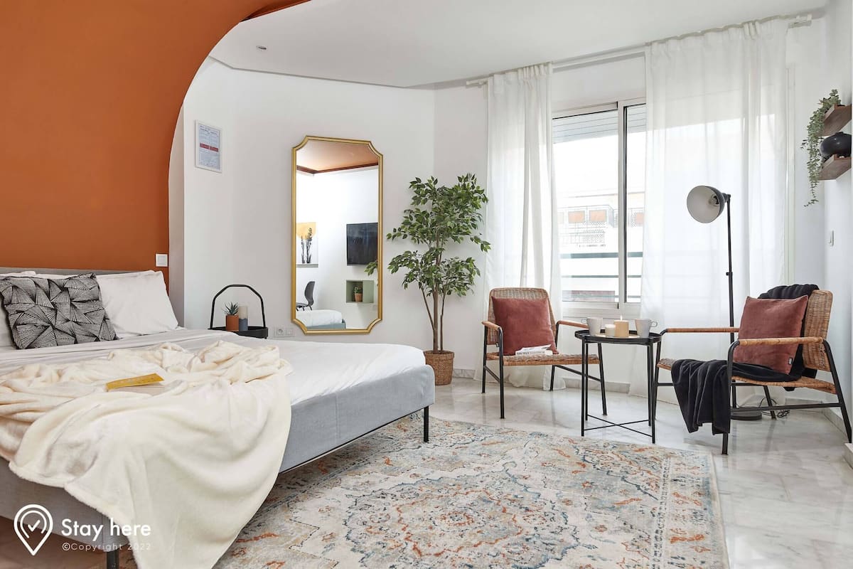 A hotel room with a bed, a large mirror, a potted plant, two chairs, a small table, and a window with curtains.
