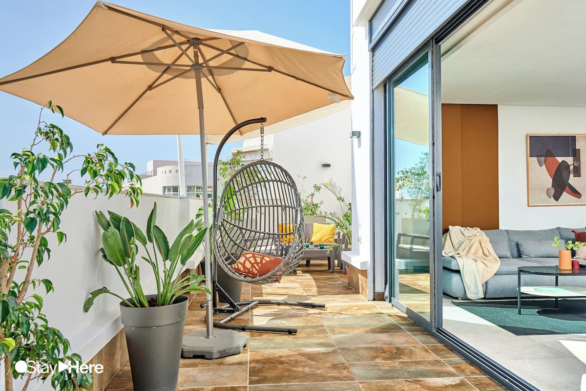 A modern outdoor patio with a swing chair, a large umbrella, and potted plants.