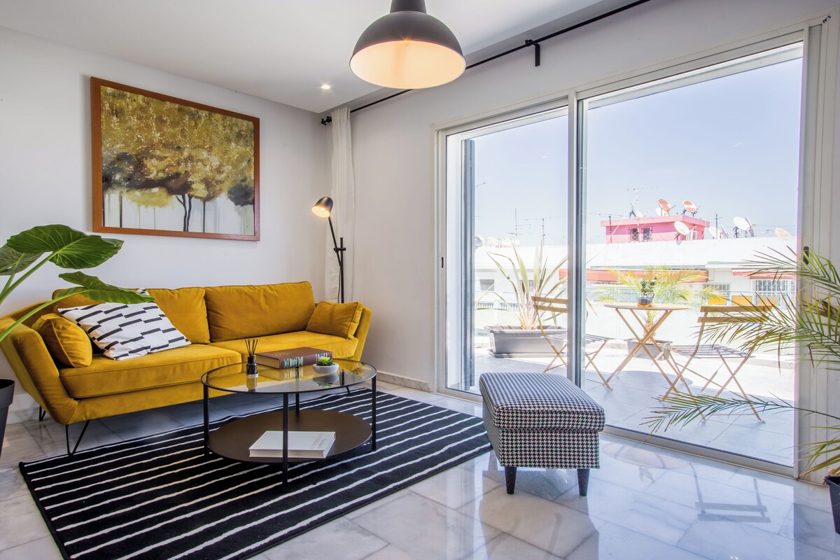 A modern living room with a yellow sofa, a glass coffee table, a black and white striped rug, and large windows offering a view of the city.