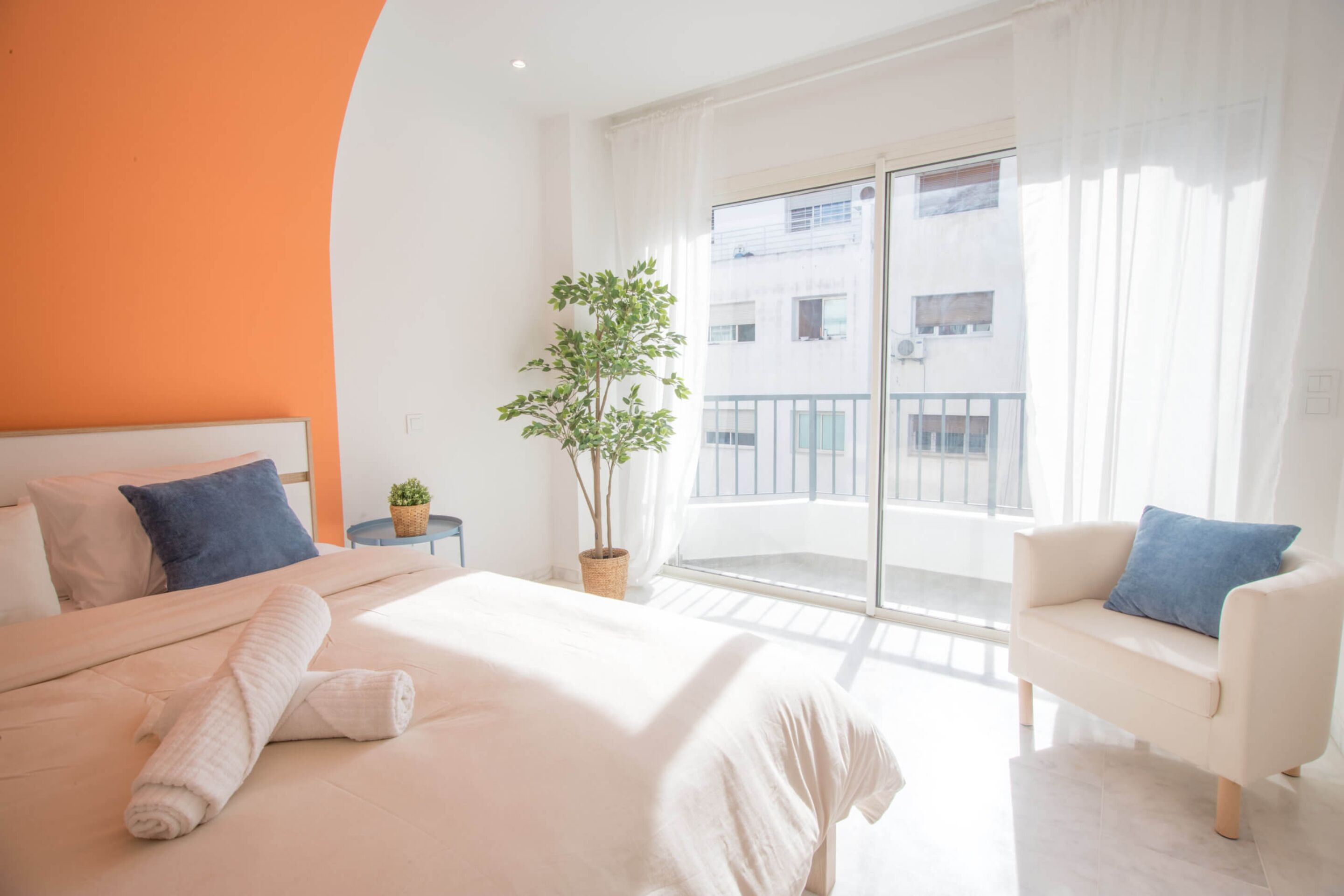 Une chambre d’hôtel avec un lit, un canapé, une plante en pot et un balcon donnant sur des immeubles.