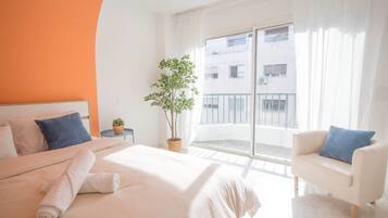 Une chambre d’hôtel avec un lit, un canapé, une plante en pot et un balcon donnant sur des immeubles.