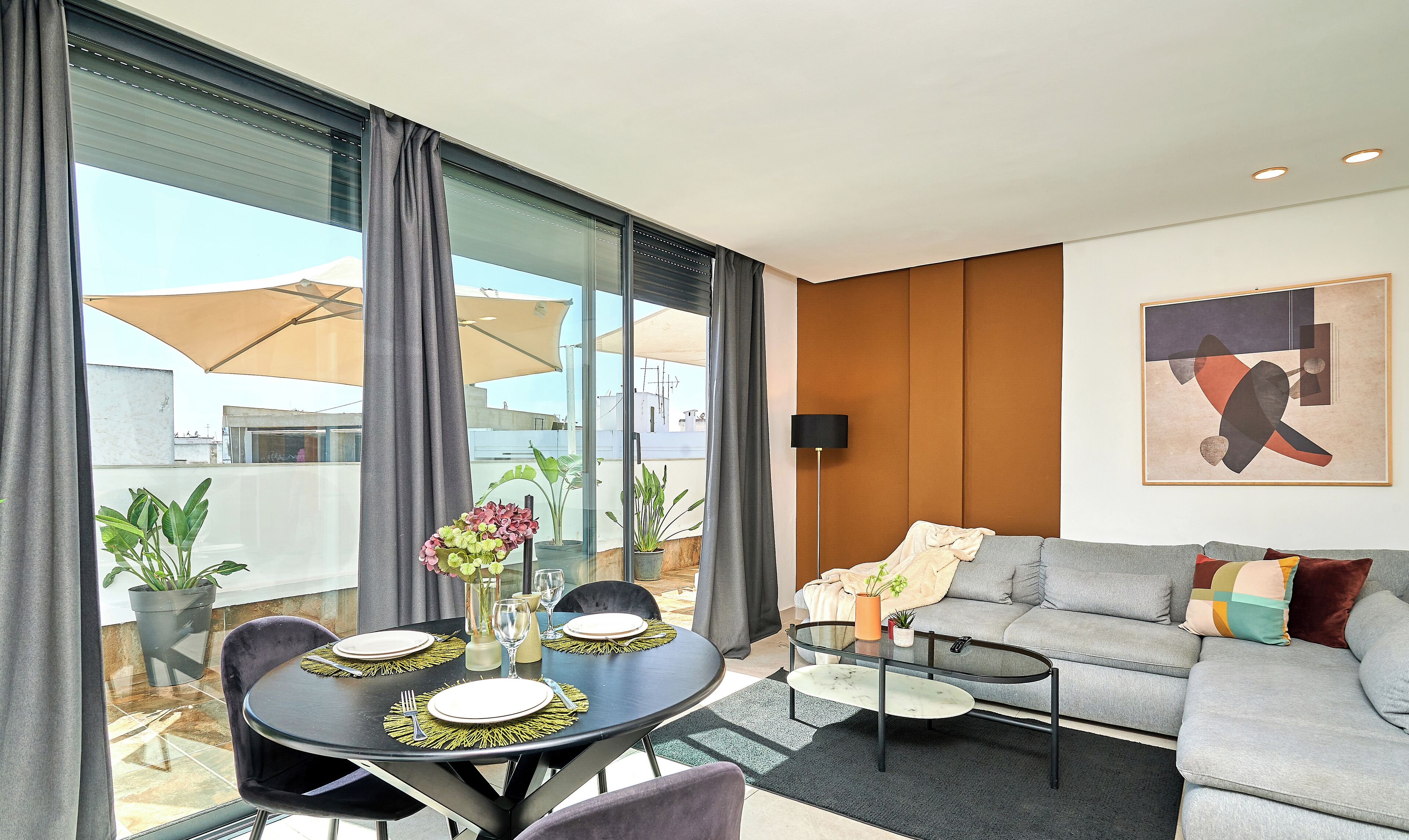 A modern living room with a grey sofa, a glass dining table set for two, and a large window with curtains.