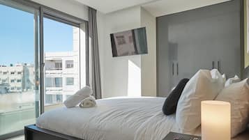 Une chambre d’hôtel moderne dotée d’un grand lit, d’un téléviseur fixé au mur et offrant une vue sur la ville grâce à de grandes fenêtres.