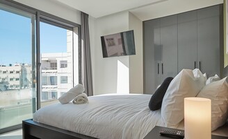 Una habitación de hotel moderna con una cama grande, un televisor montado en la pared y vistas de la ciudad a través de amplios ventanales.