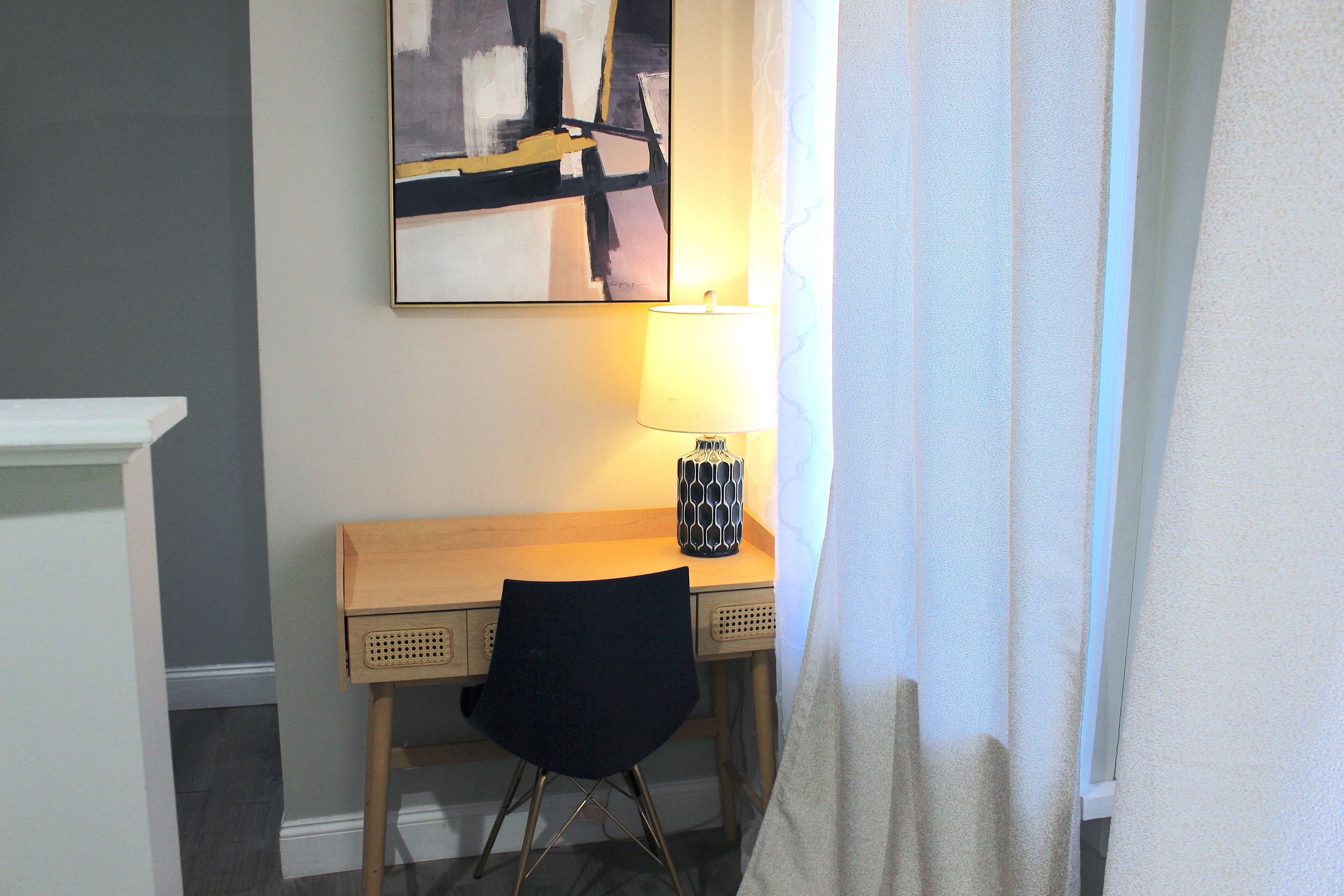 A hotel room with a wooden desk, a black chair, a lamp, and a painting on the wall.