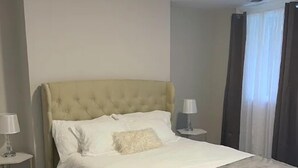 Quarto com cabeceira estofada, roupa de cama branca, manta cinza e mesa de cabeceira com abajur.