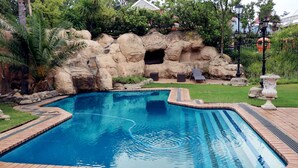 Une piscine rectangulaire bordée de briques, entourée d’un jardin aménagé avec des palmiers et une formation rocheuse.