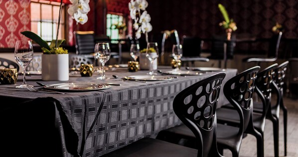 Una mesa de comedor dispuesta con platos, cubiertos y copas de vino, rodeada de sillas negras con cortes circulares.