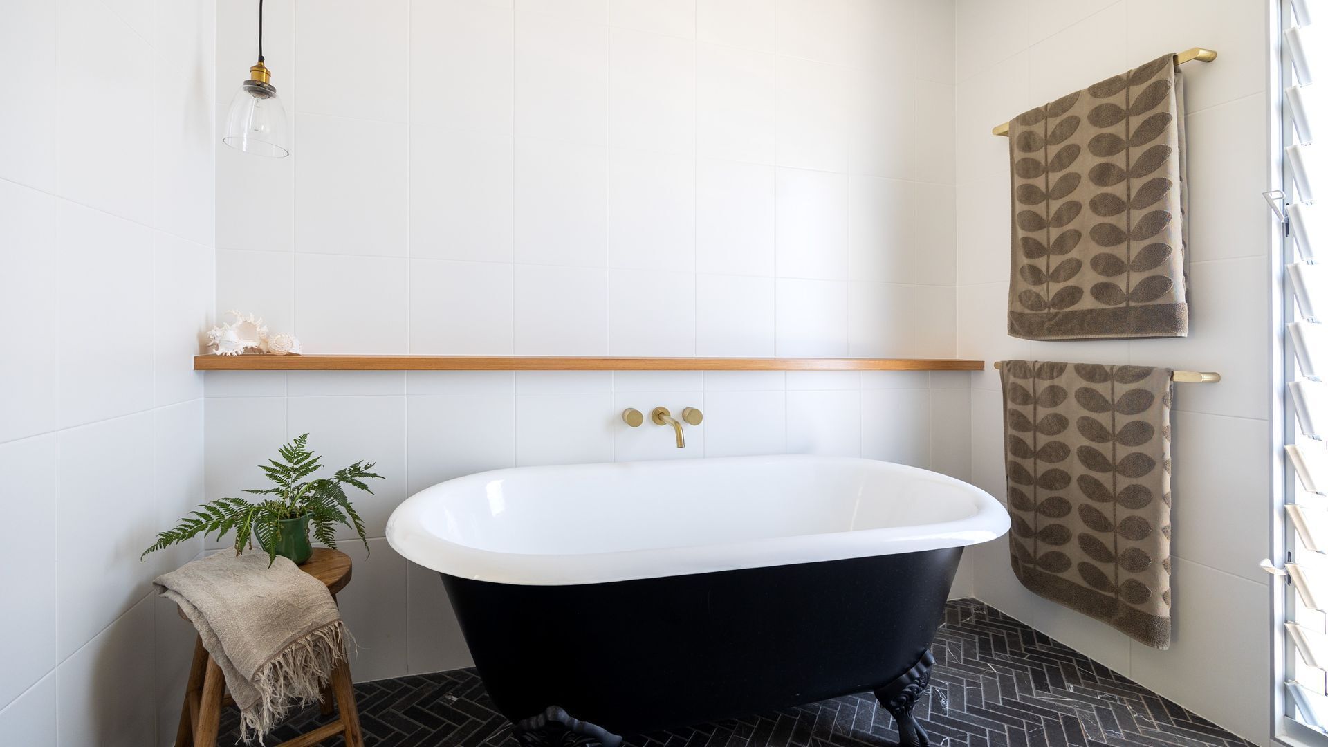 Une salle de bain avec une baignoire sur pieds, un porte-serviettes orné de serviettes à motifs, une petite plante posée sur un tabouret et une étagère avec des objets décoratifs.