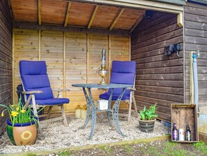 A wooden cabin with two blue chairs and a table, a potted plant, and a small wooden box with bottles.