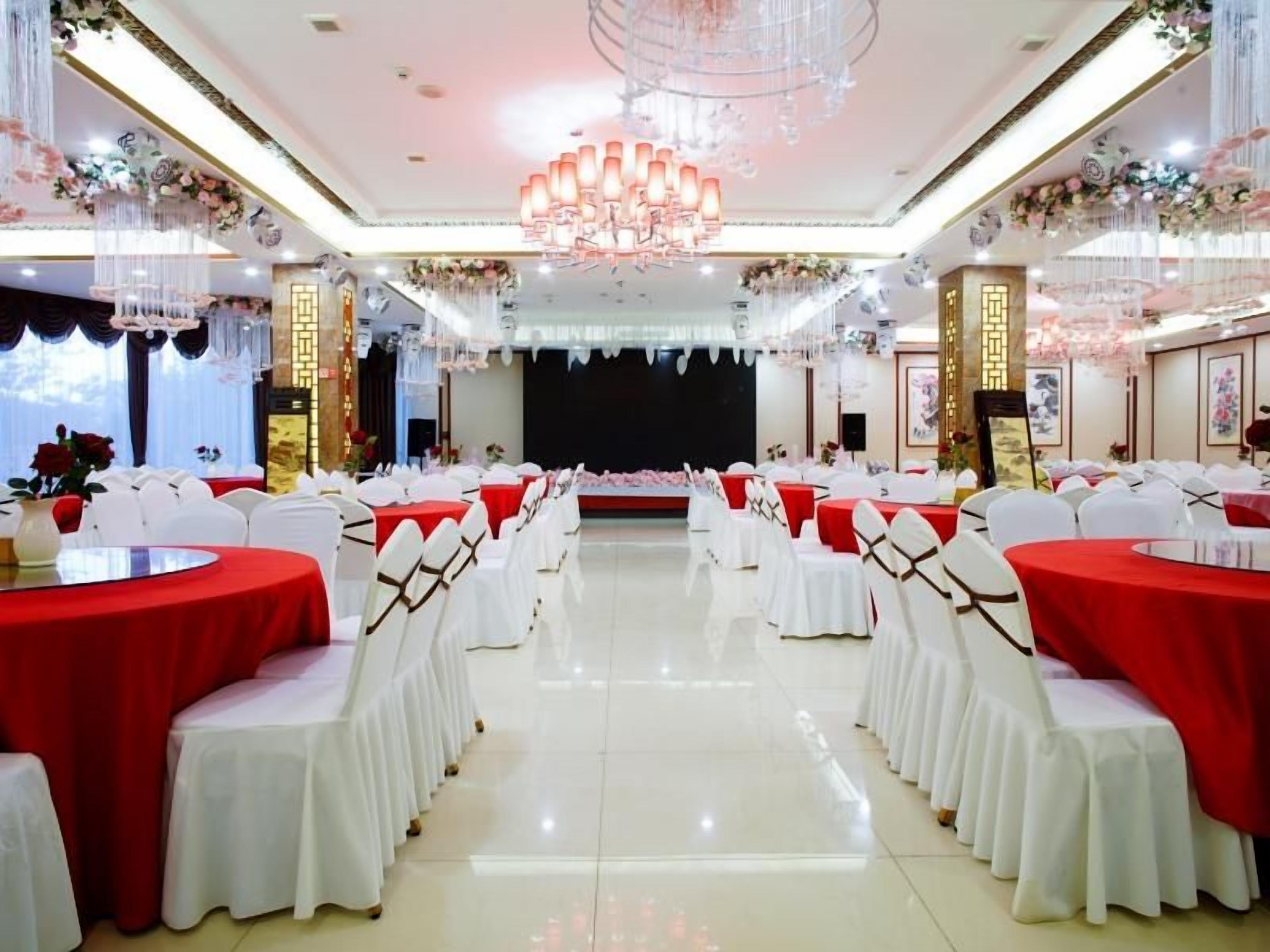 Une salle de banquet avec des tables rondes recouvertes de nappes rouges et des chaises blanches, une scène à l’avant et des compositions florales décoratives.