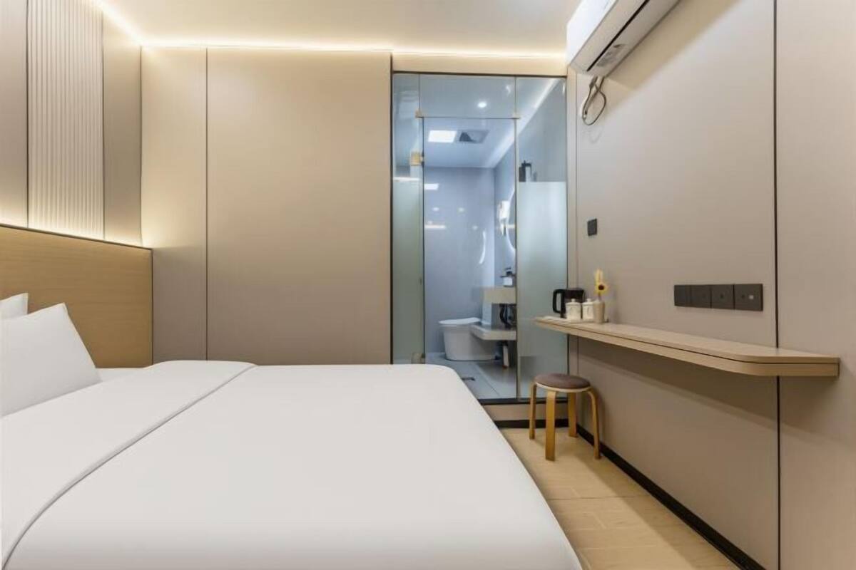 Une chambre d’hôtel moderne avec un grand lit, un petit tabouret, une coiffeuse avec un miroir et une étagère contenant des articles de toilette.