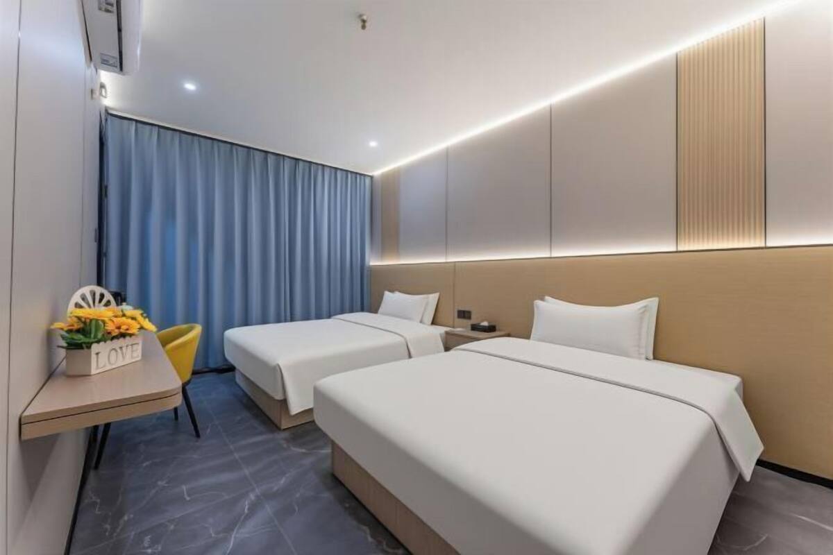 Une chambre d’hôtel avec deux lits, un bureau avec une chaise, une petite table ornée d’une décoration représentant le mot « AMOUR », et un rideau bleu.
