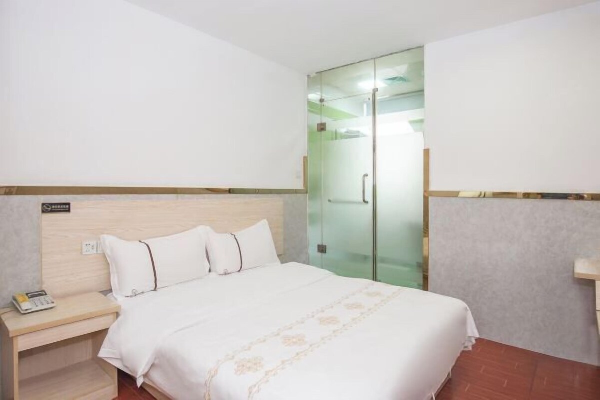 A hotel room with a bed, a bedside table with a phone, and a glass door leading to a shower area.