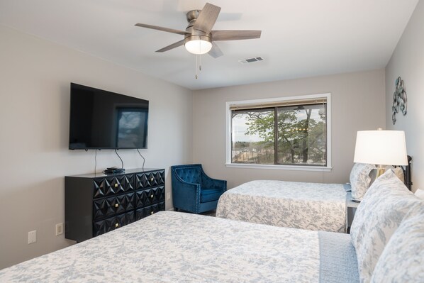 Un dormitorio con dos camas, ventilador de techo, televisor, ventana con vistas y cómoda.