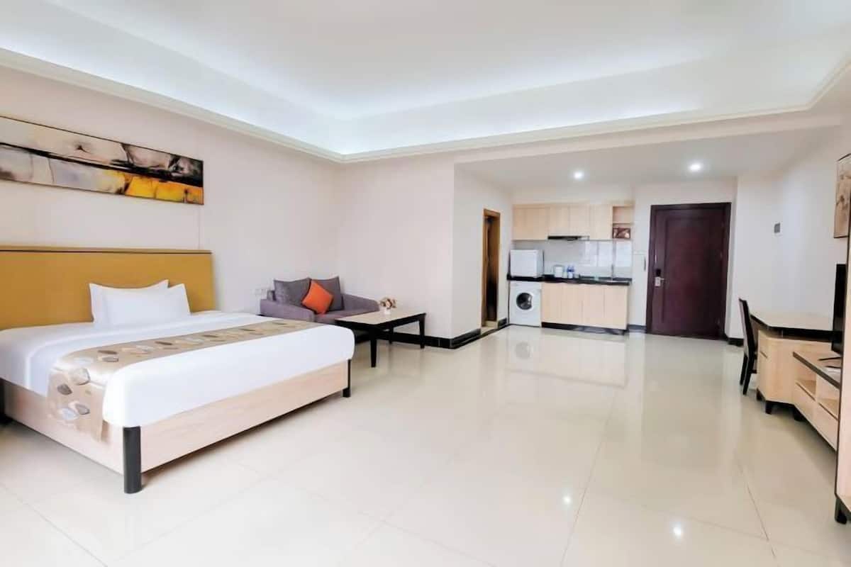 A modern hotel room with a bed, a sofa, a small table, and a kitchenette in the background.