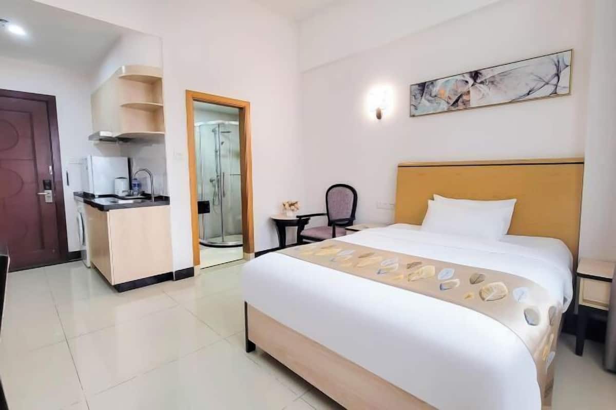 A hotel room with a bed, a small desk, a chair, a kitchenette, and a shower area.