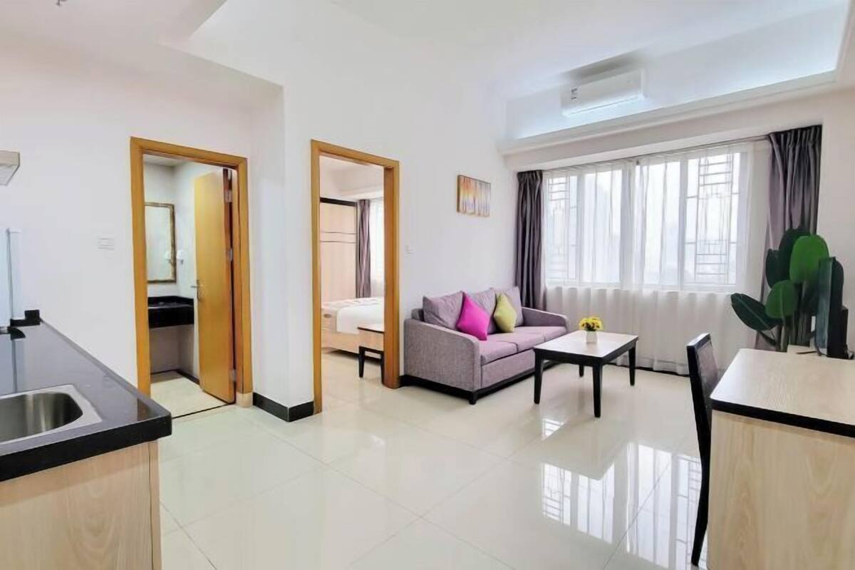 A modern living room with a sofa, coffee table, and dining table. There is a kitchen area with a sink and cabinets.