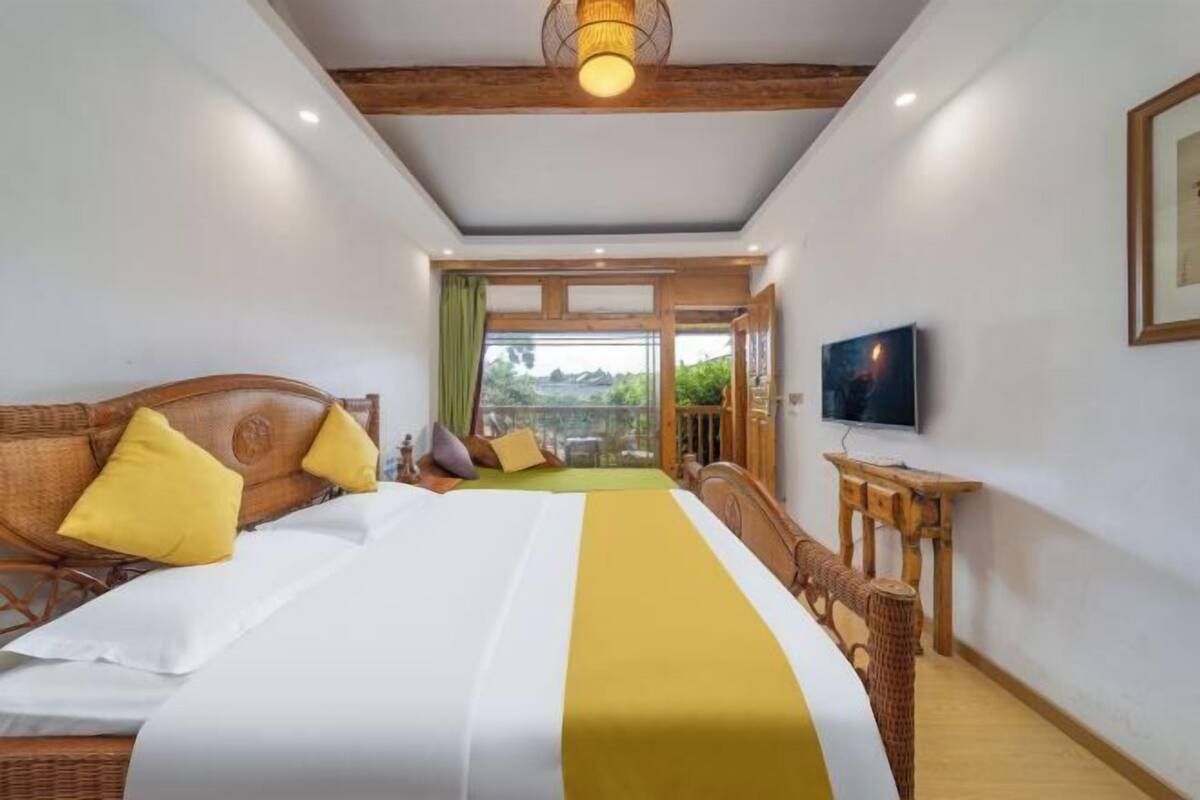 A hotel room with a large bed, a television, a wicker chair, and a balcony with a view.