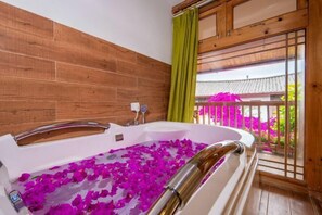 A bathtub filled with water and rose petals, wooden walls, a green curtain, and a view of a traditional building through a window. - Yuhuai Renjia Inn (Lijiang)