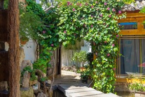 A garden pathway with a wooden bridge, surrounded by greenery and a yellow building. - Yuhuai Renjia Inn (Lijiang)