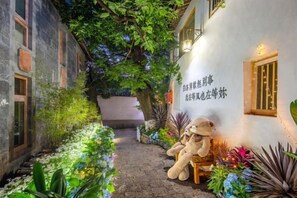 A narrow alleyway with a white building on the right, a teddy bear on a bench, and various plants. - Yuhuai Renjia Inn (Lijiang)