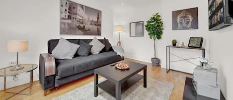 A modern living room with a grey sofa, a coffee table, a glass table, and a TV. There are wall art pieces and a plant.