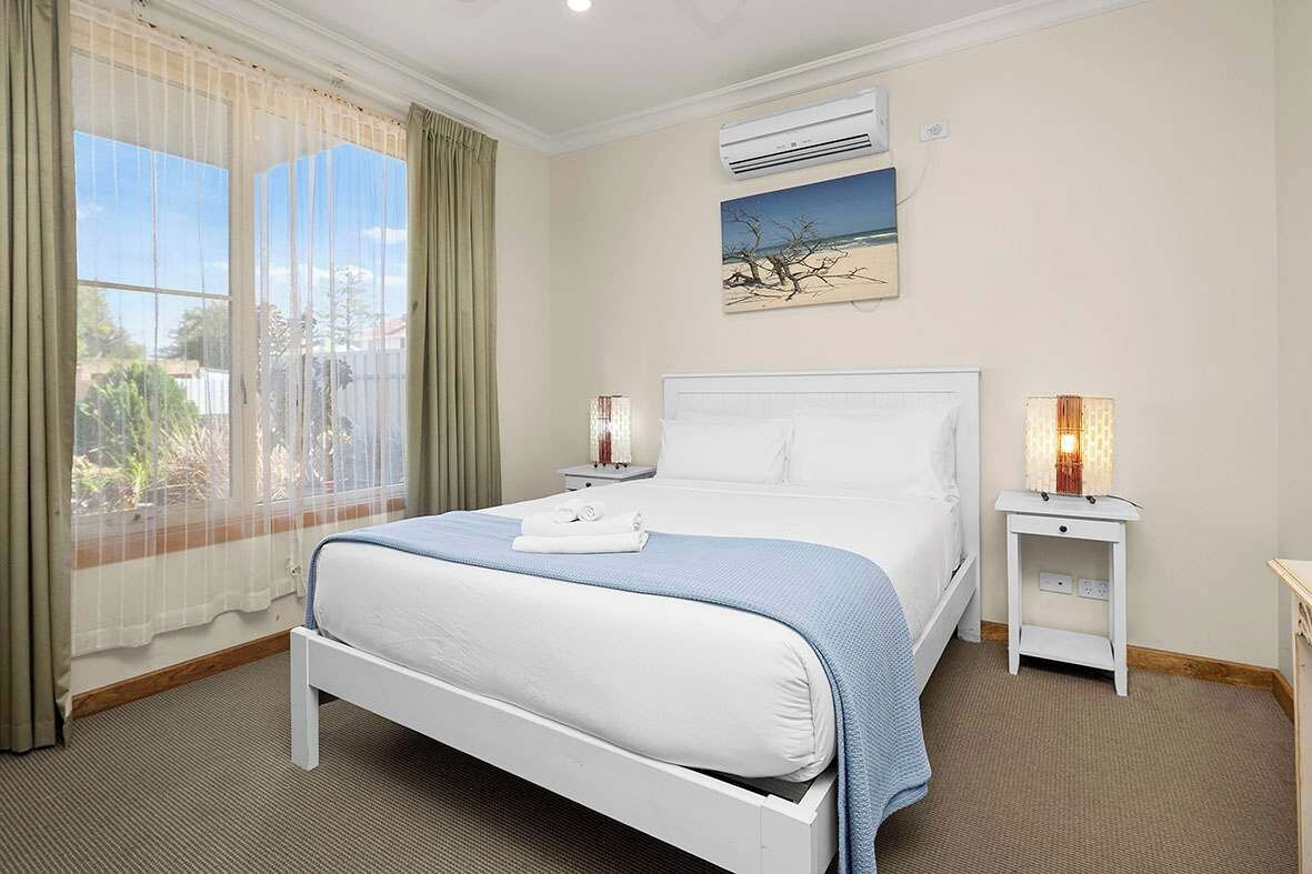 A hotel room with a bed, two bedside tables, an air conditioning unit, a window with curtains, and a painting on the wall.