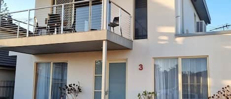 A modern two-story building with a balcony, a door marked with the number 3, and potted plants in the foreground.