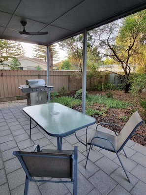 An outdoor patio with a table and chairs, a grill, and a ceiling fan. - Sandrift by the beach (Largs North)
