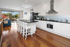 A modern kitchen with white cabinetry, a central island with bar stools, a gas stove, and a large window overlooking a backyard. - Sandrift by the beach (Largs North)
