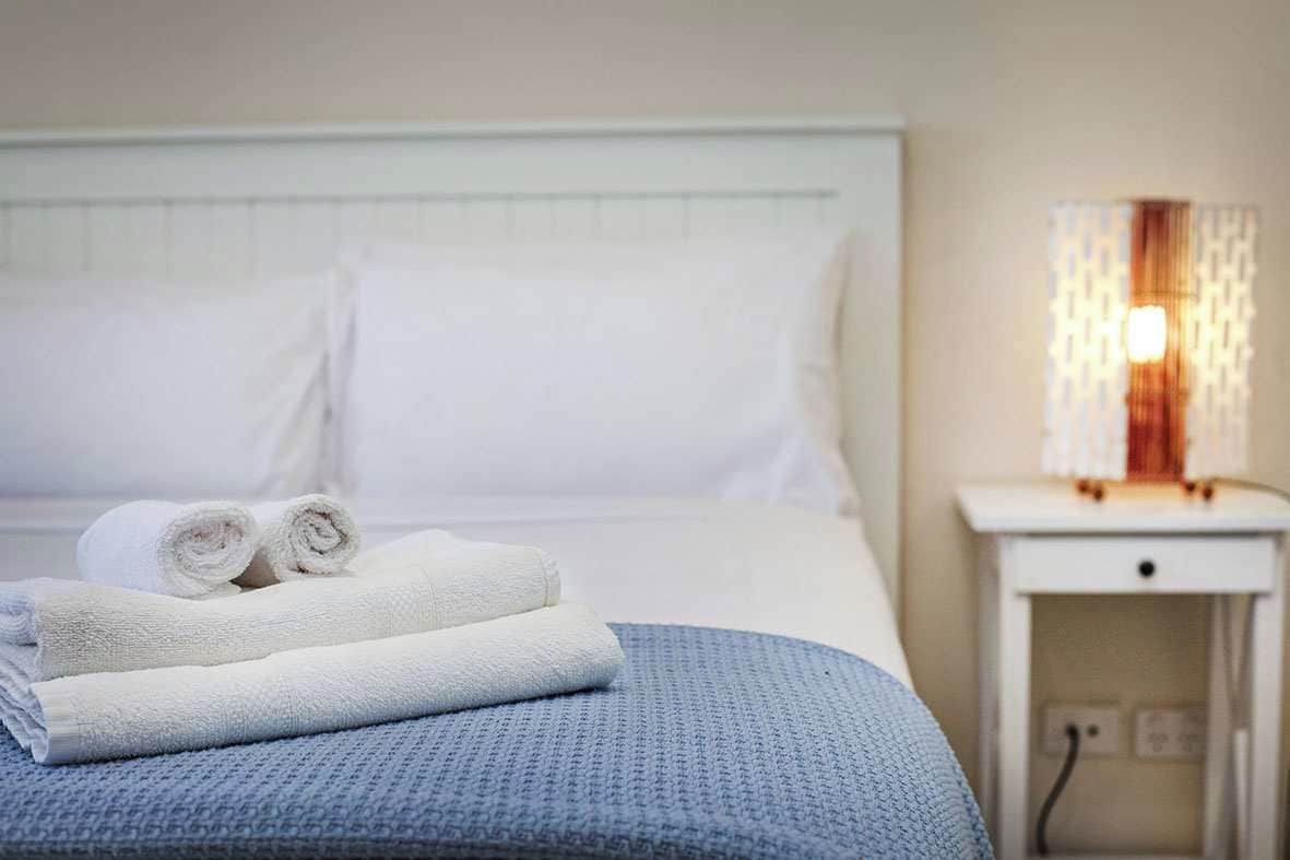 A neatly made bed with rolled white towels and a blue blanket, a white bedside table with a lamp, and a white wall.